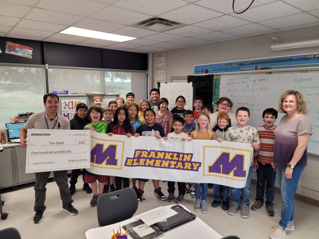 Photo from Tim Odell's 5th grade classroom at Franklin Elementary after he won the Bridgestone Class of Quarter Grant. Photo is staff member, students, Bridgestone rep holding a banner. 